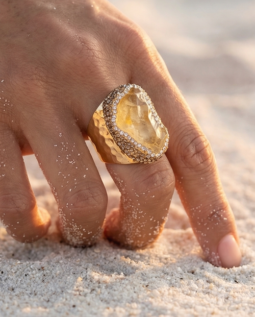 Hand wearing a gold ring with a rough diamond cut stone on a sandy background
